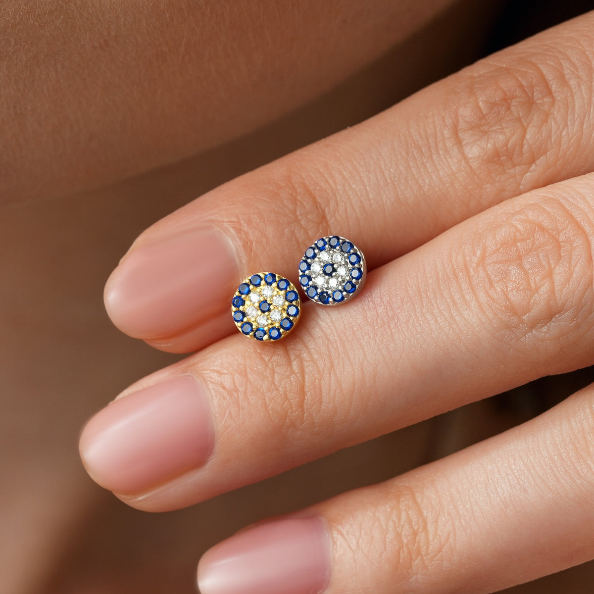 Gold vermeil and sterling silver evil eye stud earrings with blue and clear stones, shown on fingertips.