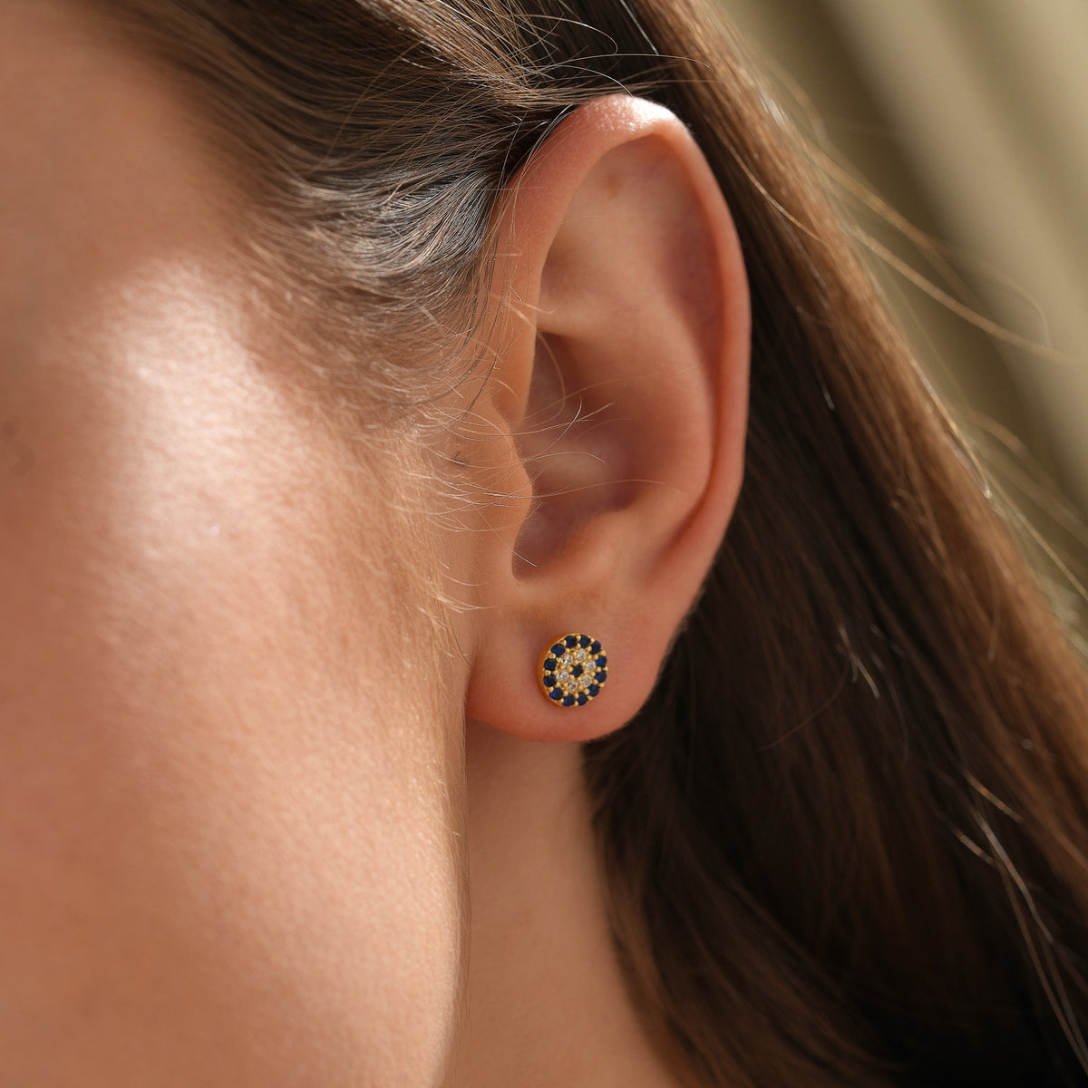 Gold vermeil evil eye stud earring with blue and clear stones, shown close up on the ear.