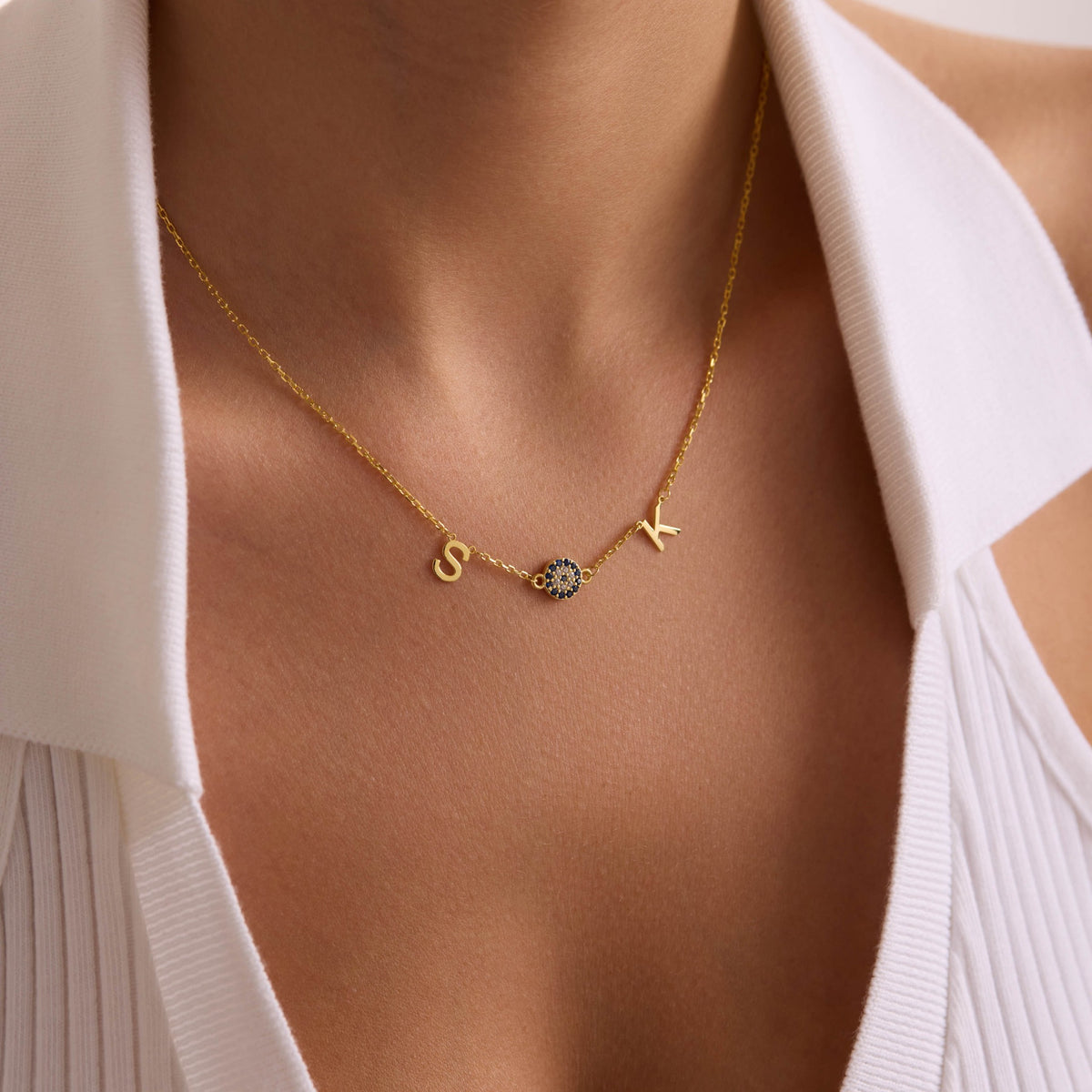 Personalised gold initial necklace with S and K letters and a blue evil eye, worn at the centre of the neckline.