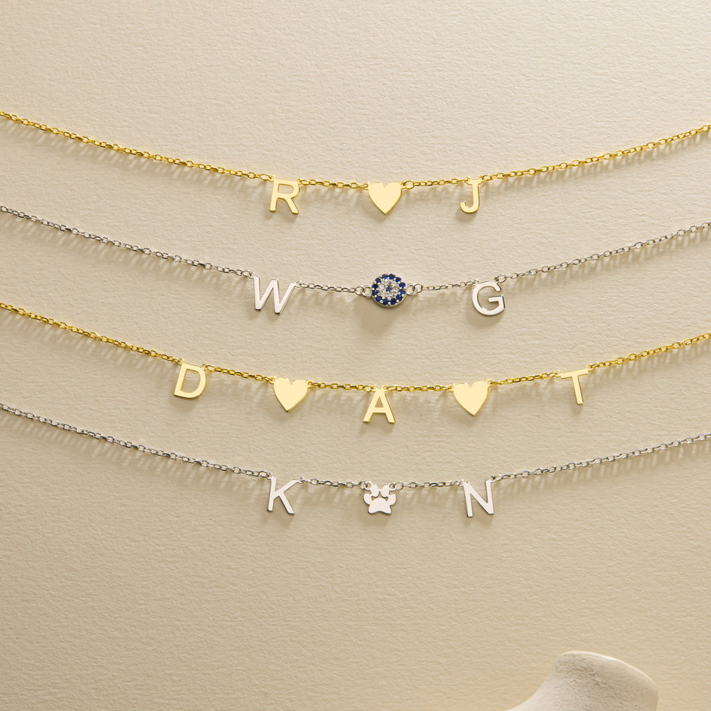 Personalised gold and silver initial necklaces with heart, blue evil eye and paw symbols arranged centrally on a cream background.