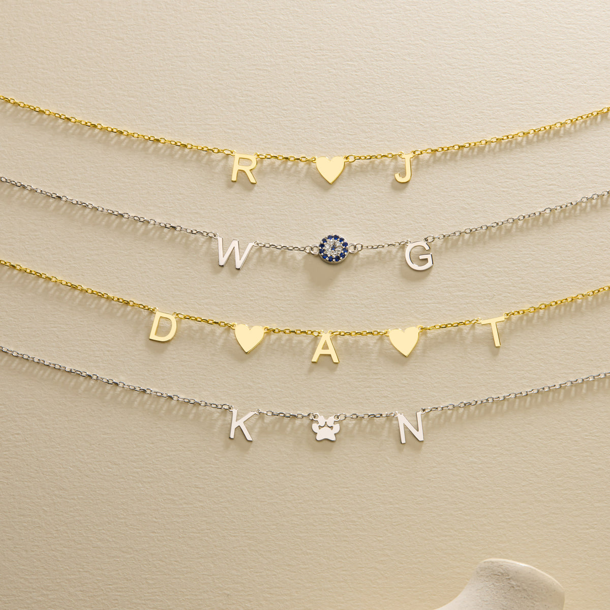 Personalised gold and silver initial necklaces with heart, blue evil eye and paw symbols arranged centrally on a cream background.
