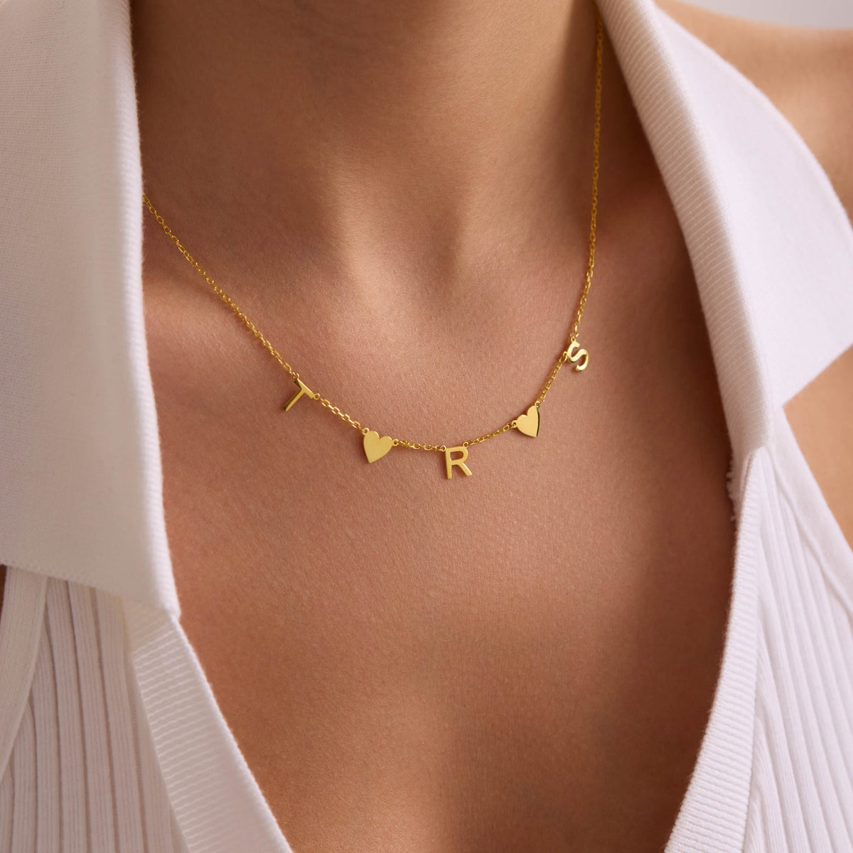 Gold multiple initial necklace with T, R and S letters and two heart charms, worn centrally.