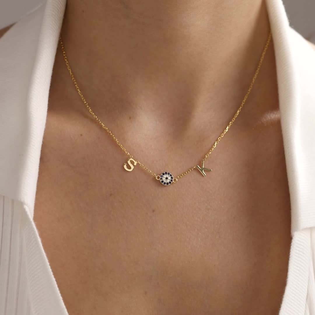 Sideways Multiple Initial & Evil Eye Necklace in gold vermeil, showing personalised initials with a blue evil eye and a heart charm design worn at the neckline.
